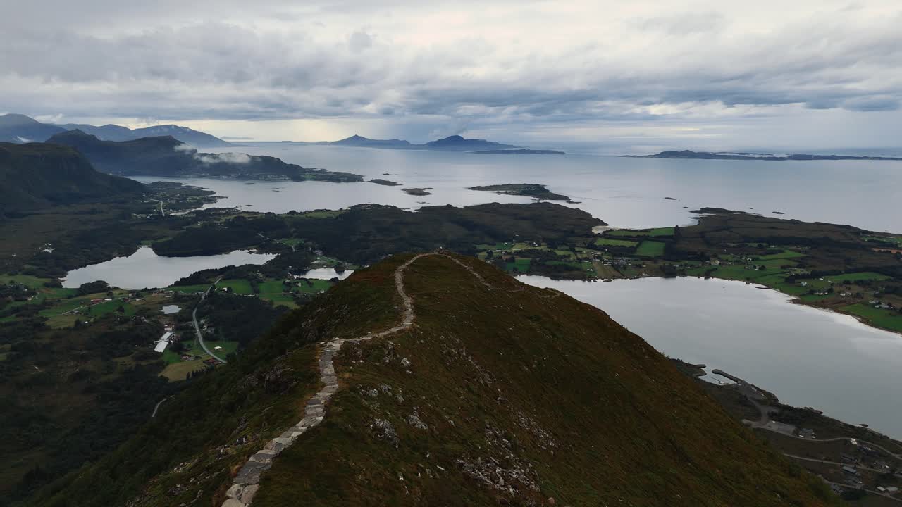 Steiniger Wanderweg auf schmalem Gebirgsgrat mit grüner Vegetation und dramatischer Fjordlandschaft darunter, Norwegen