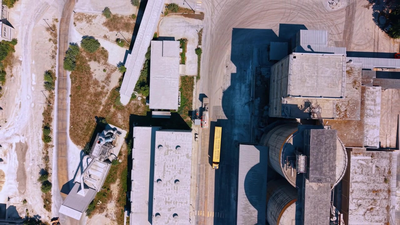 Aerial top down view of industrial plant with vehicles. Long shadows stretch across concrete structures and paved ground around a yellow truck during daylight.