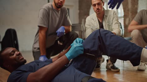 close-up shooting: a black male doctor in a blue medical uniform demonstrates the tightening of a medical tourniquet on his leg for public. practical trainings on first medical aid. course of a medic