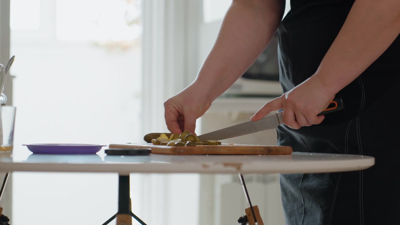 Close-up of person wearing dark apron slicing cooked vegetables on wooden cutting board beside backlit kitchen table, showing active knife motion, finger positioning, and casual home cooking