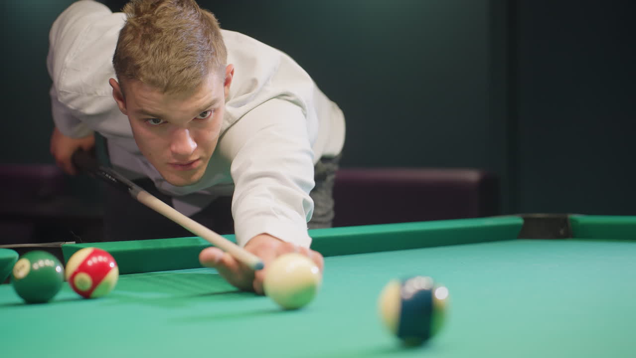 Front view of player in white shirt with serious face, leaning over green pool table in dimly lit room. Gripping cue stick with focus, ready to strike, displaying intense concentration and precision