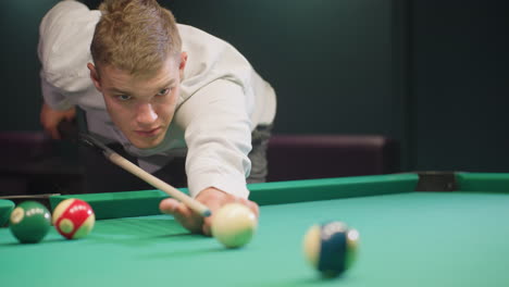 front view of player in white shirt with serious face, leaning over green pool table in dimly lit room. gripping cue stick with focus, ready to strike, displaying intense concentration and precision