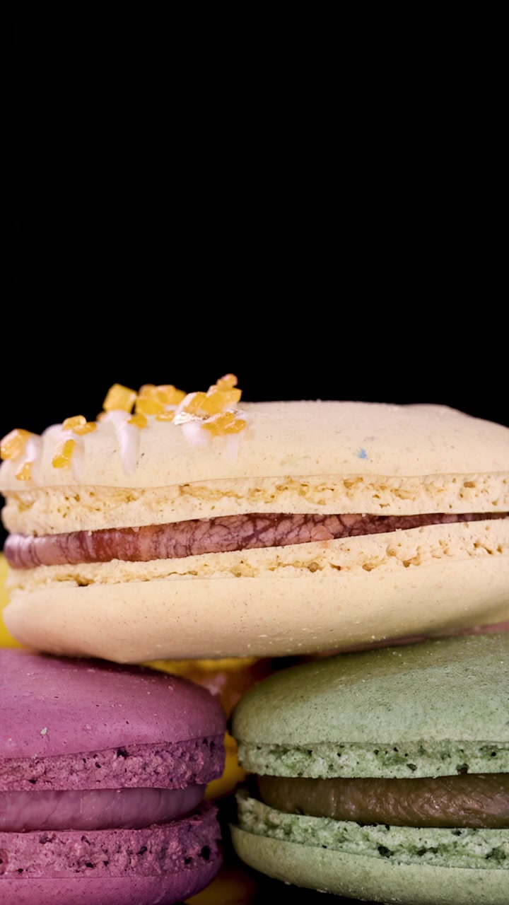 A hand places a decorated macaron atop a stack of assorted colorful macarons, shot in close-up with dramatic lighting and a black background