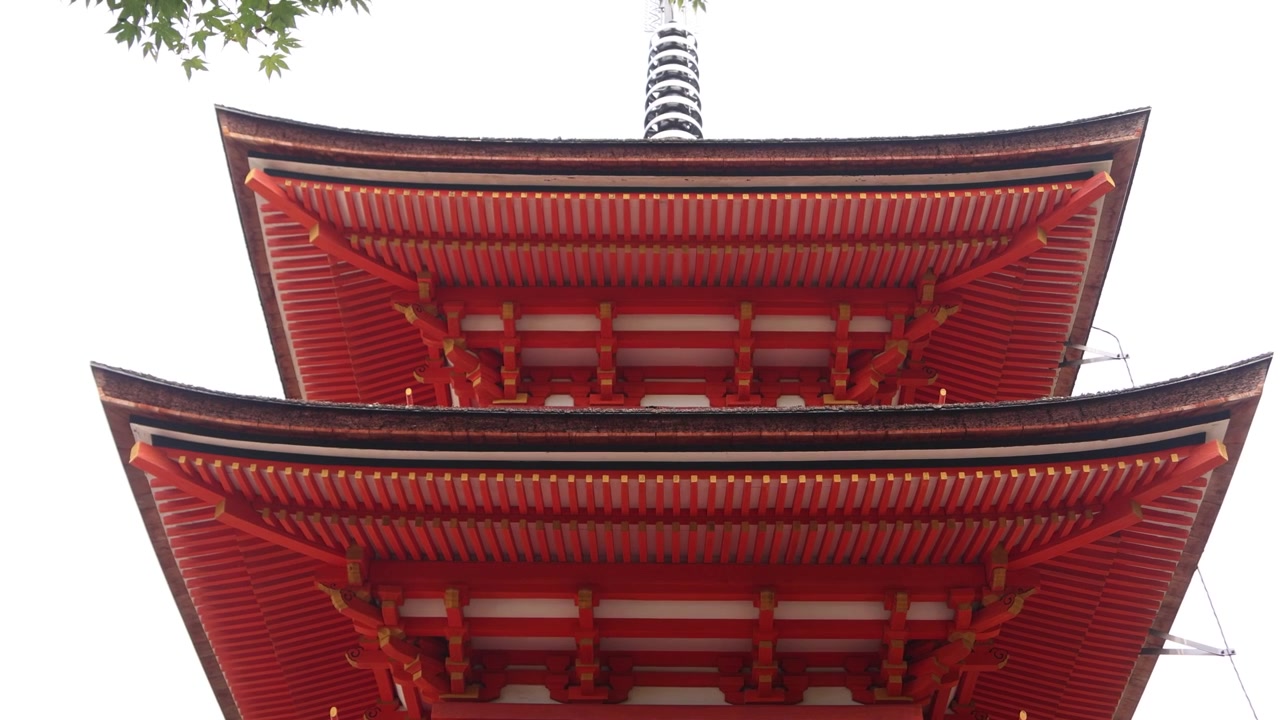 Opadgående udsigt til Kiyomizu-deras cinnoberrøde pagode i Kyoto, der fremhæver dens lagdelte træetager, elegante taglinjer og traditionelle buddhistiske design mod Higashiyamas lyse himmel.