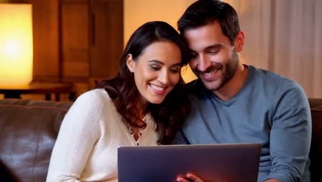 a happy young couple sits together on a couch at home, using a laptop.