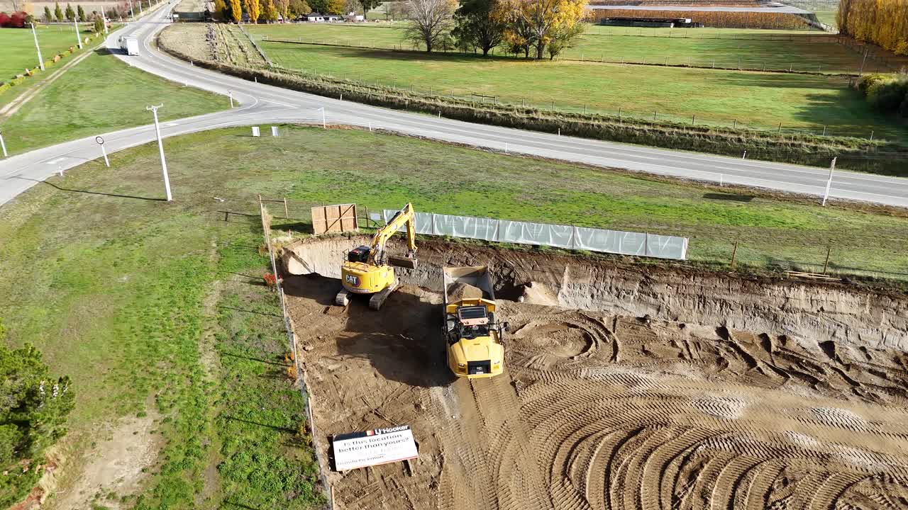 Drone footage captures an excavator and dump truck working on a construction site in Cromwell, New Zealand, under clear daylight