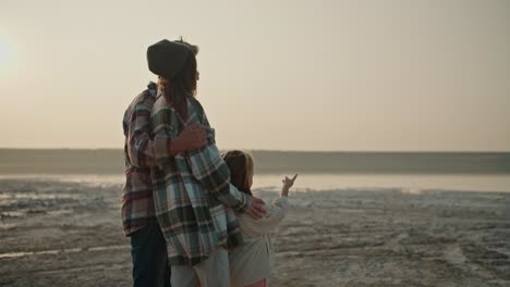 Rear-view-of-a-happy-brunette-man-with-gray-hair-in-a-checkered-shirt-stands-and-hugs-his-wife-and-daughter-during-his-vacation-outside-the-city-on-a-deserted-seashore-in-a-summer-evening