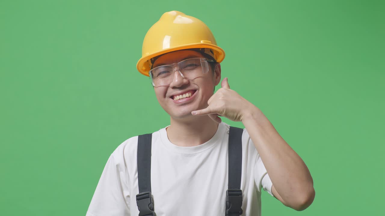 Close Up Of Asian Man Worker Wearing Goggles And Safety Helmet Smiling And Making Call Me Gesture To Camera While Standing In The Green Screen Background Studio