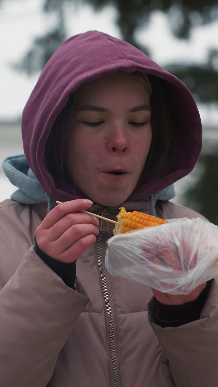 dama con chaqueta de invierno comiendo maíz en la calle al aire libre, sosteniendo maíz envuelto en plástico, disfrutando de comida callejera, clima frío, comida de invierno casual, chaqueta con capucha, entorno de parque urbano