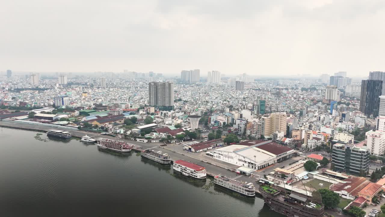Aerial view of Ho Chi Minh City, Vietnam, featuring a densely packed urban skyline with high-rise buildings, riverside docks, and moored boats along the waterfront on a cloudy day