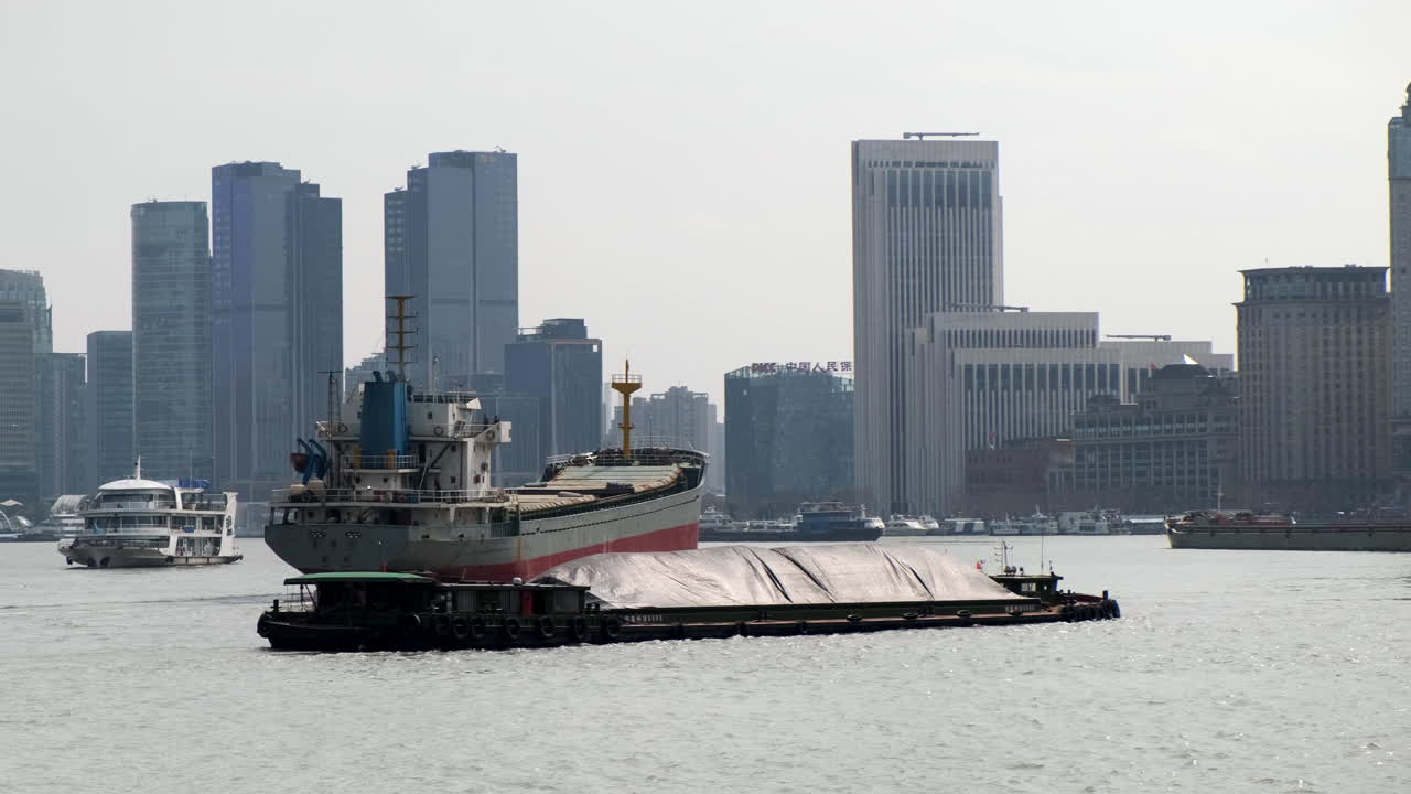 Large ships and barges navigating the bustling Huangpu River, with the Shanghai skyline in the hazy background