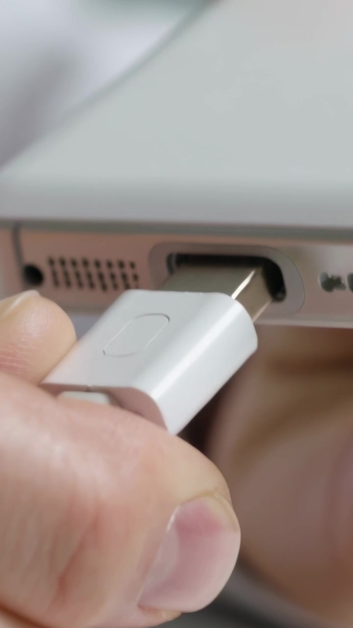 Vertical video: Holding connector, hands inserting white cable into mobile port at desk, charging