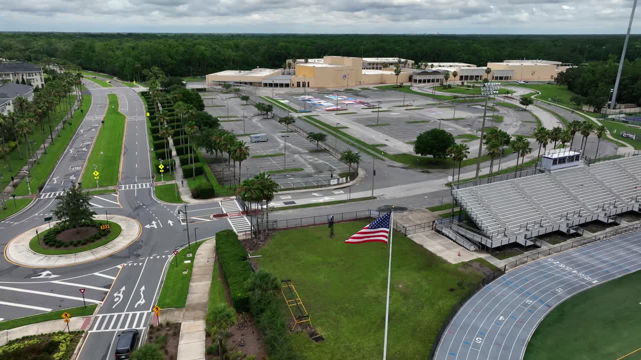 Roundabout with waving american flag and driving car at cloudy day in Celebration, Florida. Aerial wide shot. Yellow facade of Celebration High School with empty parking area. Palm trees on street.