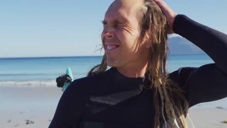 video portrait of happy caucasian male surfer with dreadlocks standing on sunny beach in wetsuit