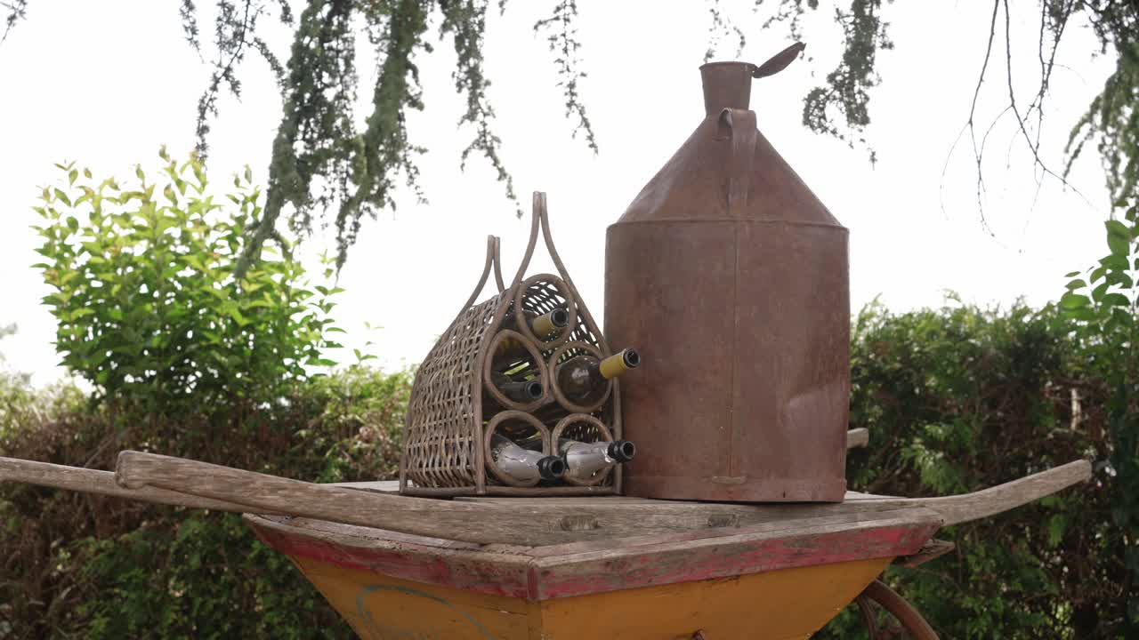 Outdoor scene with wine bottles in wicker holder and rusty metal jug on a wooden cart in garden