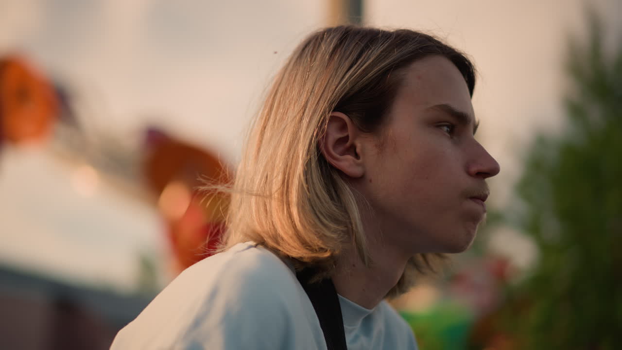 Chitarrista adolescente bianco al crepuscolo del luna park, capelli biondi lunghi fino alle spalle di profilo, espressione pensierosa, tracolla della chitarra visibile, illuminazione cinematografica calda, giostre carnevalesche sfocate e bokeh, ritratto ravvicinato