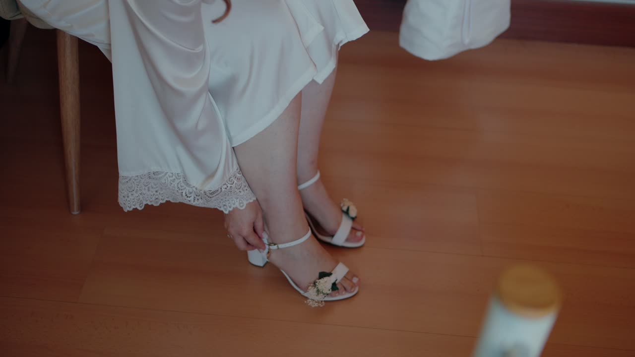 Bride in white robe fastens delicate floral white sandals on light wooden floor