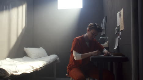 female inmate thoughtfully writing letter at desk in isolated prison cell