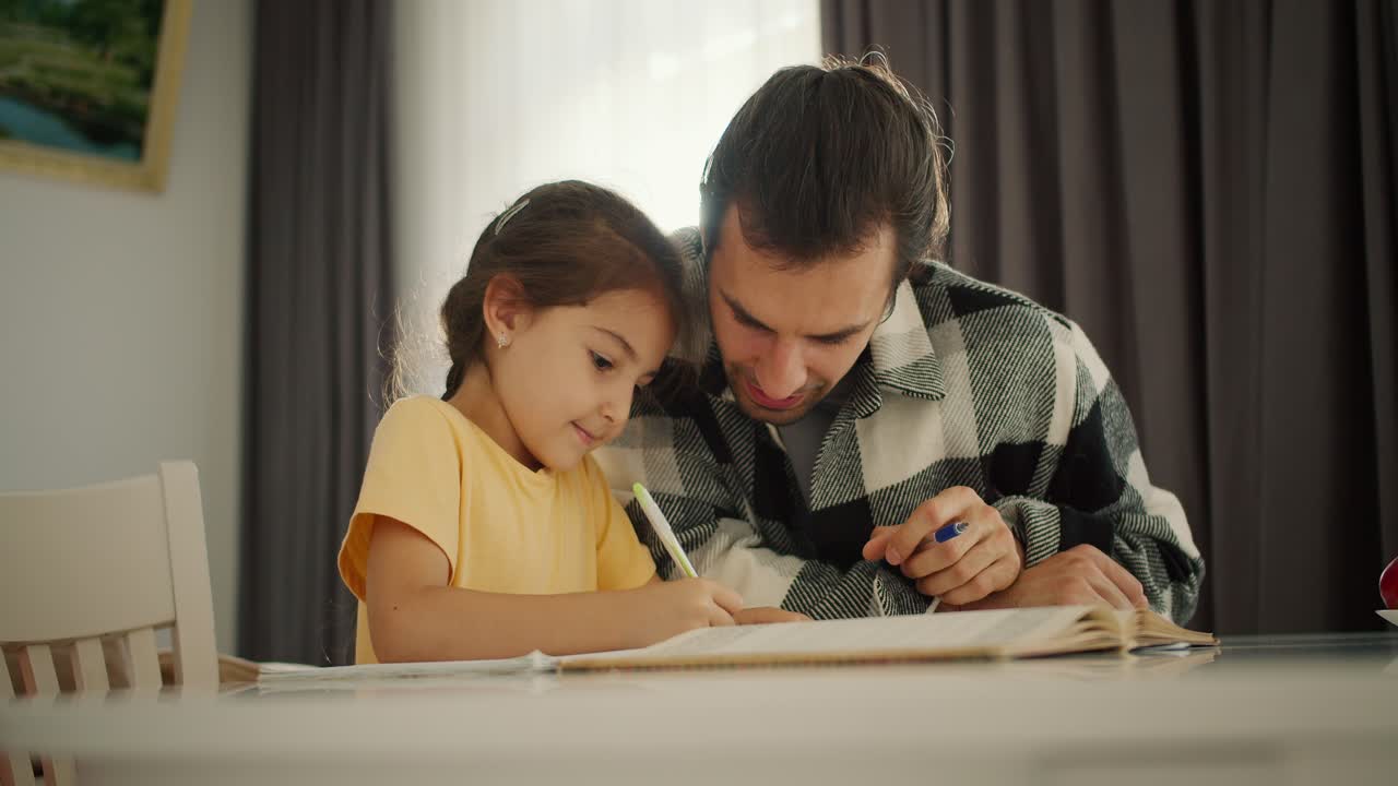 una pequeña chica morena en un vestido amarillo hace su tarea y escribe en su cuaderno junto con su padre, un hombre moreno en una camisa a cuadros en una mesa blanca en una habitación en un apartamento moderno