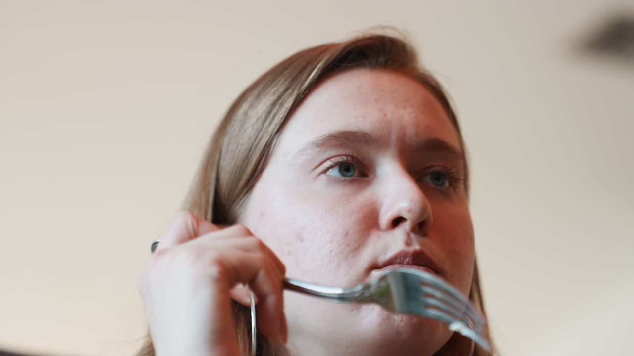 Potret jarak dekat wanita muda mencicipi salad dengan gigitan perlahan dan penuh perhatian, anting hoop dan bintik-bintik terlihat, kamera fokus pada bibir dan garpu, reaksi wajah yang ekspresif, cahaya alami yang lembut, potret jarak dekat makan malam yang intim.