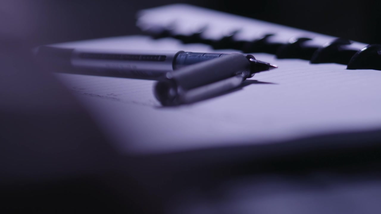 Hands typing on a keyboard in the foreground of a pen and notepad.