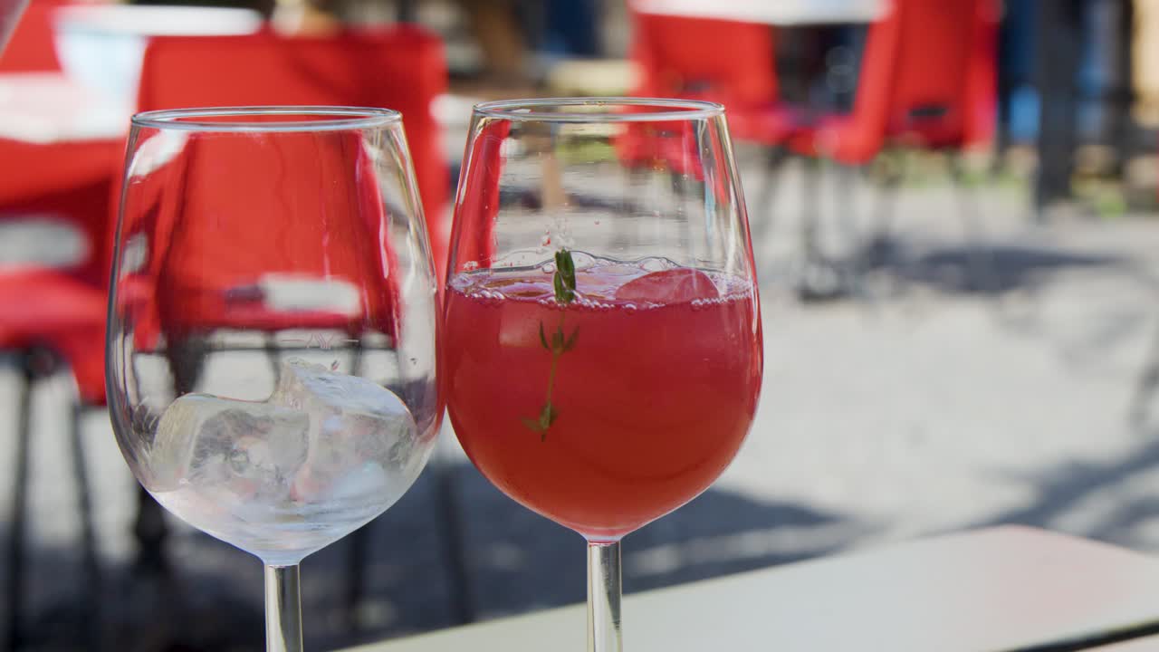 Succo di frutta rosso viene versato da una brocca in due calici da vino con ghiaccio, all'aperto in un caffè soleggiato con sedie e tavoli rossi.