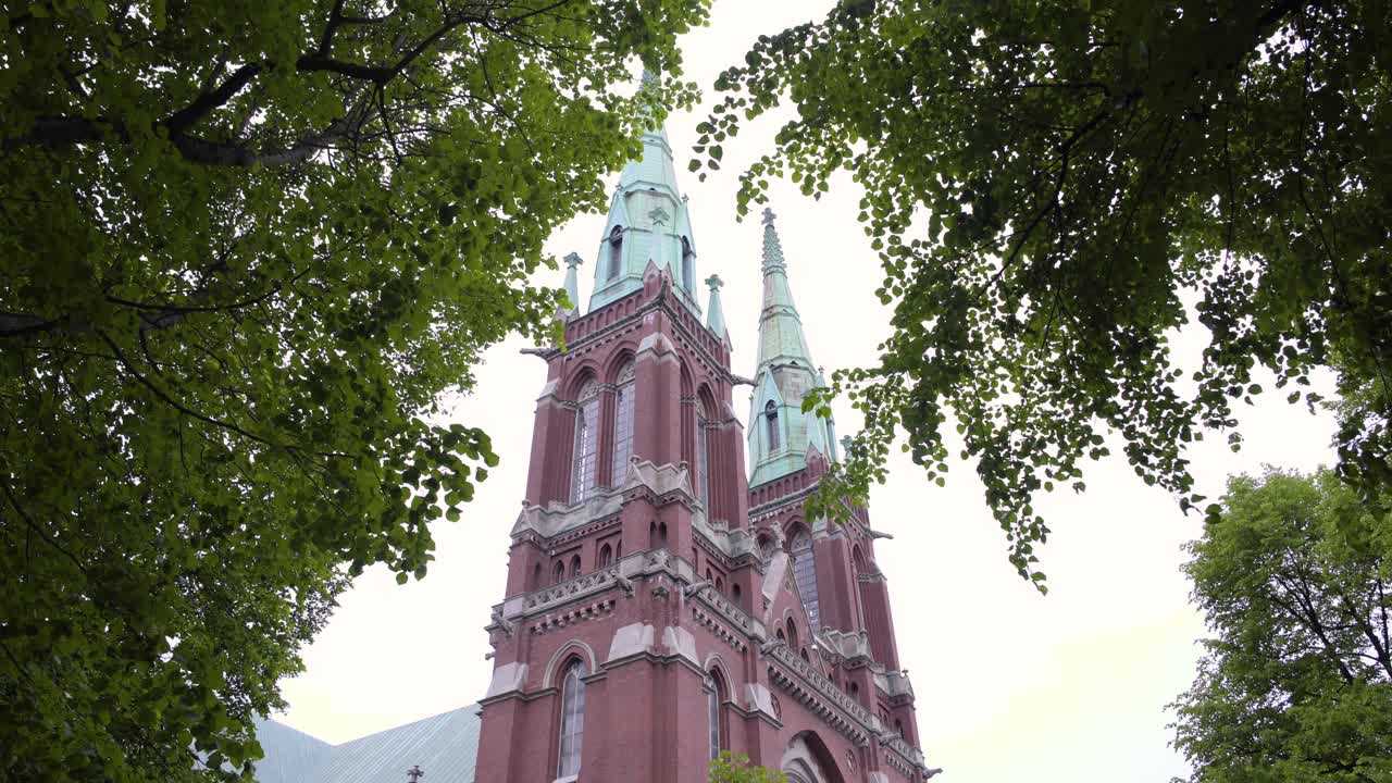 toma estática de la iglesia de san juan, filmada bajo árboles, hojas y ramas verdes, ondeando en el viento, en un día nublado de verano, en ullanlinna, helsinki, finlandia