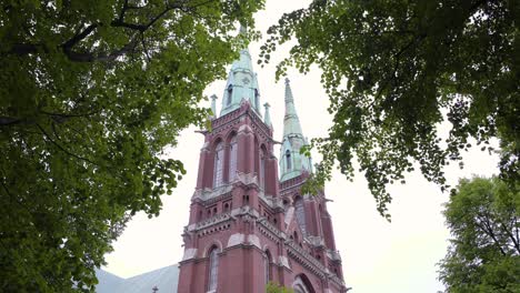 static shot of st johns church, filmed under trees, green leaves and branches, waving in the wind, on a cloudy, summer day, in ullanlinna, helsinki, finland