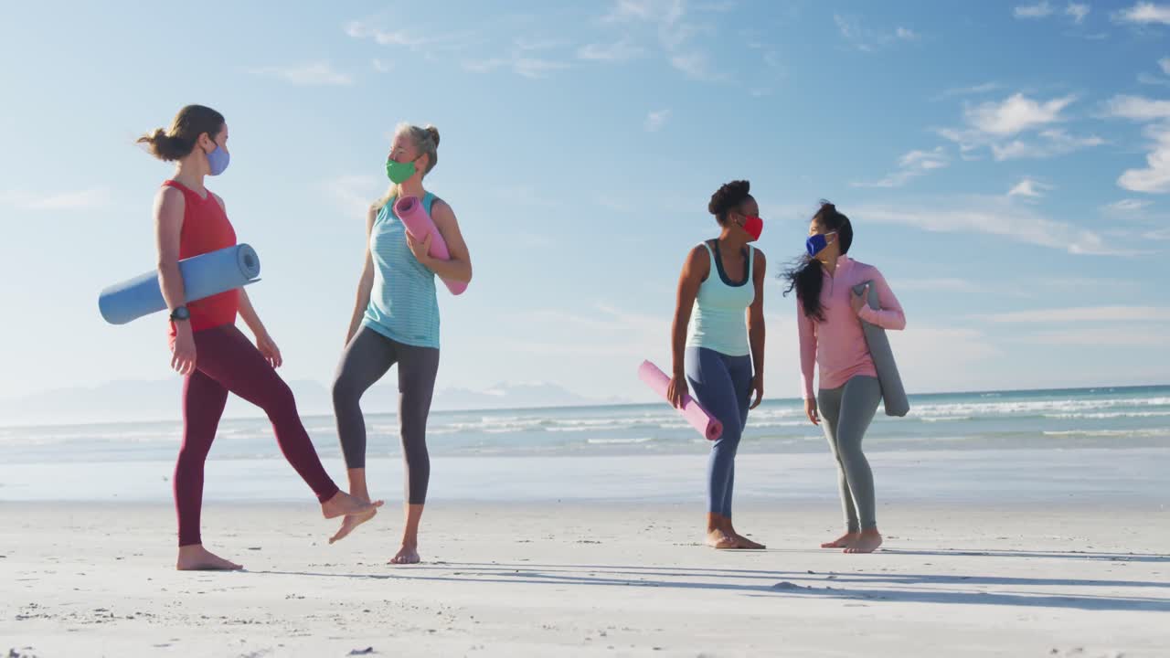Group of diverse female friends wearing face masks holding yoga mats at the beach