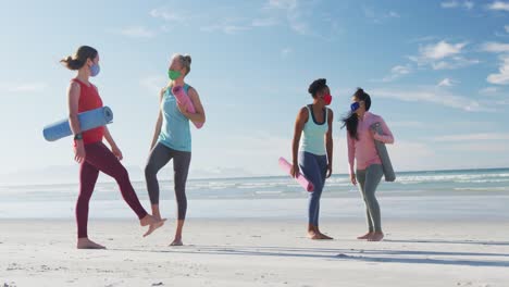 Grupo-De-Amigas-Diversas-Que-Usan-Máscaras-Faciales-Y-Colchonetas-De-Yoga-En-La-Playa