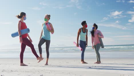 grupo de diversas amigas con máscaras faciales sosteniendo esteras de yoga en la playa