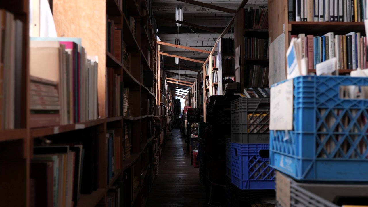 Movimento de câmera dolly pelo corredor de uma livraria passando por pilhas de livros, ângulo baixo, iluminação suave