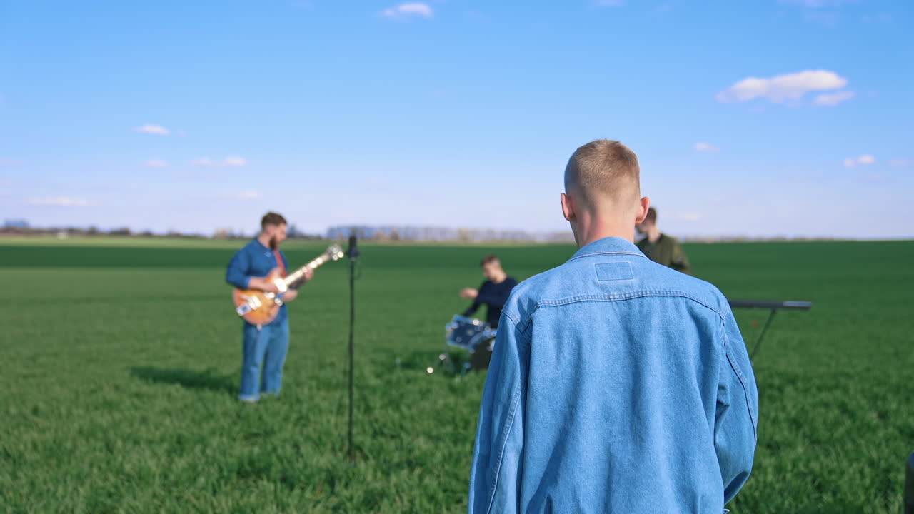 Music band on green field background. Young men playing musical instruments outdoors. Man singer coming to a microphone.
