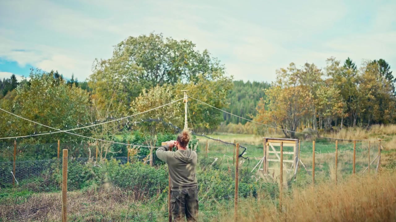 Caucasian Man Outdoors Tying Ropes In A Rustic Fence With Forested Background. Wide Shot