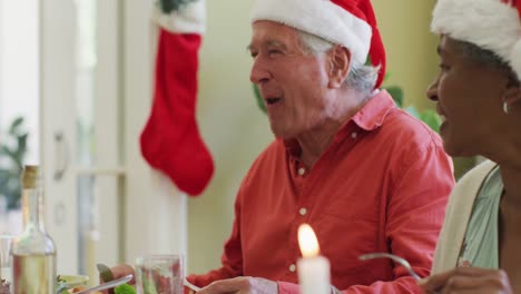 Diverse-senior-man-and-woman-in-santa-hats-listening-and-laughing-at-christmas-dinner-table-at-home