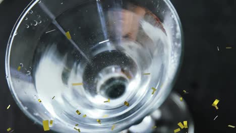 golden confetti falling over close up of olives falling into cocktail glass against black background