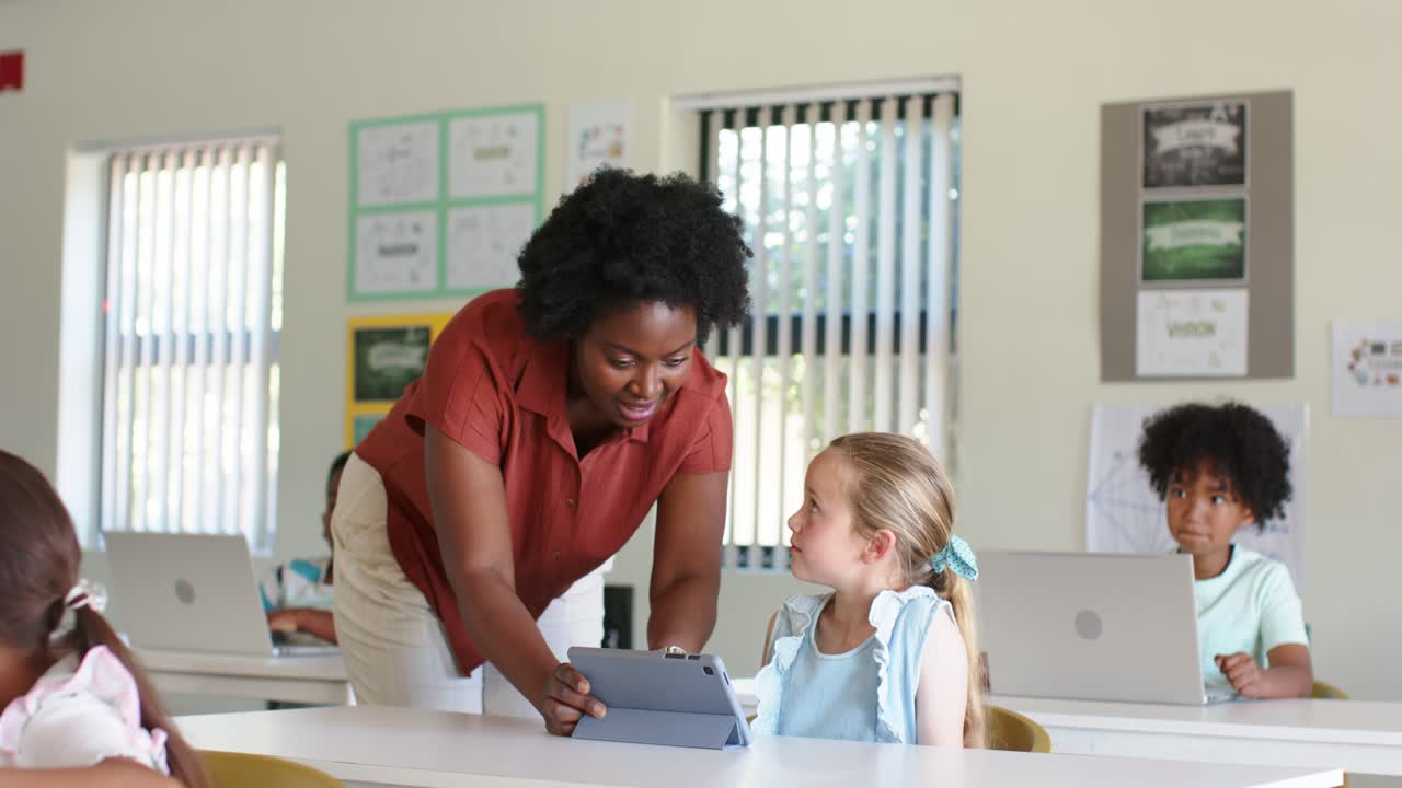 African American woman teacher noticing youth girl needing help, leaning over tablet in classroom
