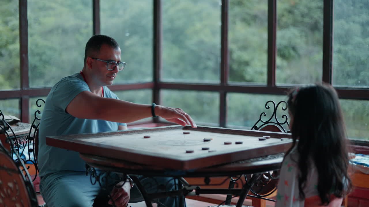 A peaceful moment of a father and daughter enjoying carrom at a restaurant table with greenery visible through large windows