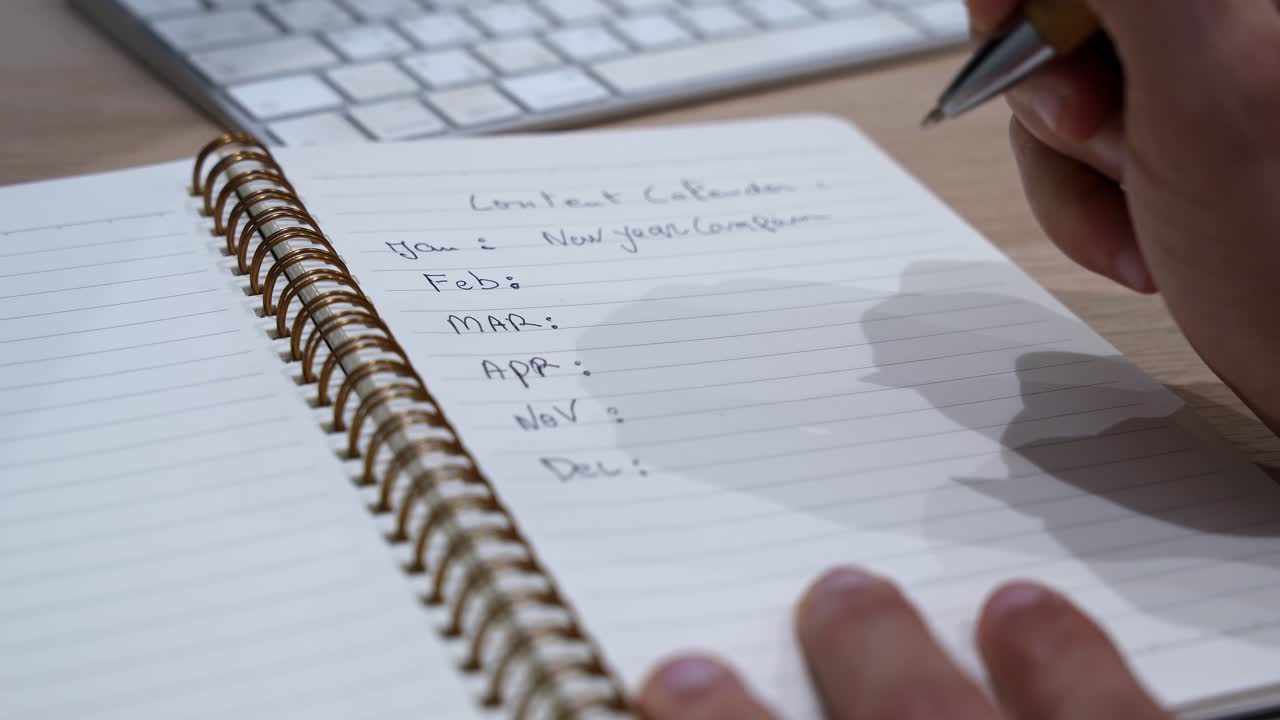 Close-up of a hand writing a content calendar in a spiral notebook on an office desk with keyboard, captured in soft indoor light with shallow depth of field