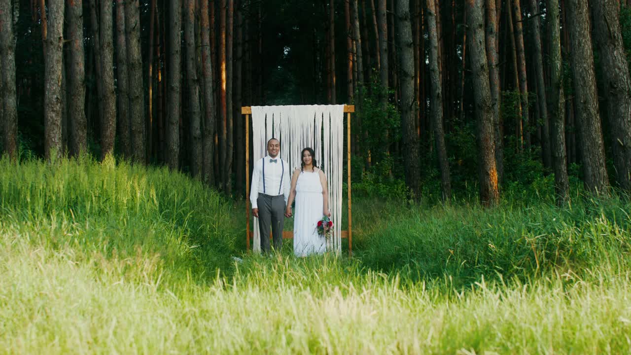 пара стоит под деревянной аркой в лесу во время свадебной церемонии