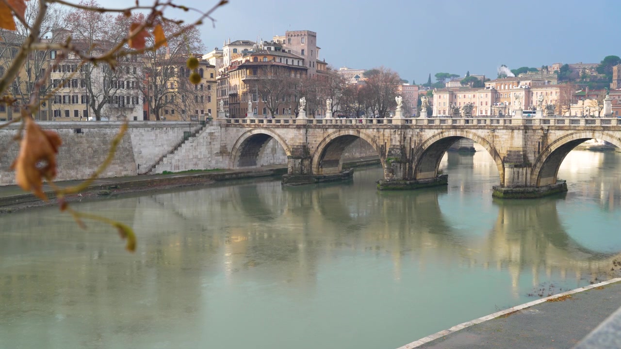 Ponte Sant'Angelo attraversa il fiume Tevere in una grigia giornata invernale, con alberi spogli e vecchi edifici di Roma che costeggiano il lungofiume.
