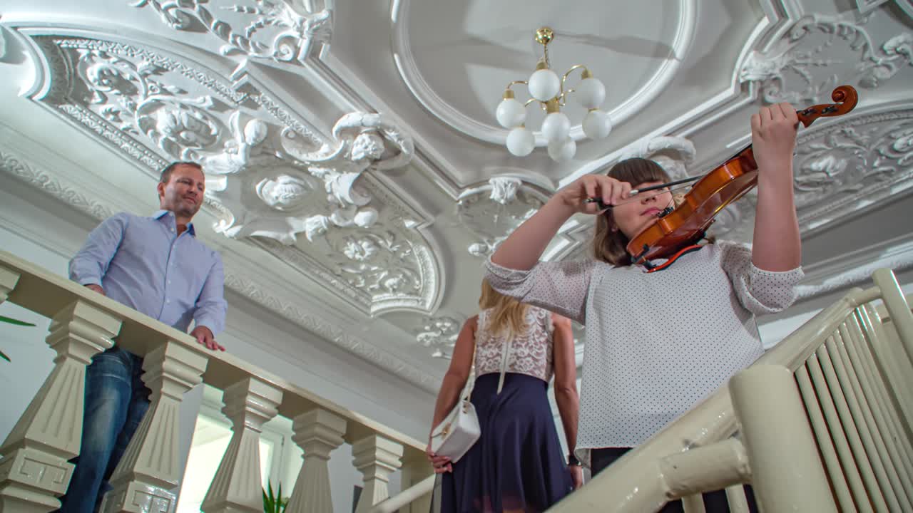Woman plays violin on staircase in old house with stucco and old lamp on the ceiling. A man listens to her and a woman walks past her up the stairs to the man. Low angle zoom in and out