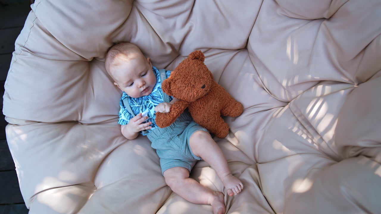 Lovely infant in blue shirt and shorts lies on the sofa outdoors. Little baby boy holding a teddy. Top view.