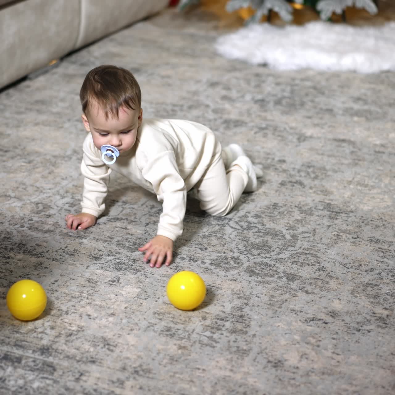 Crawling Caucasian toddler plays with balls on the floor. Black domestic cat runs near baby. Christmas tree at backdrop