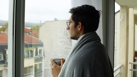 young man staring at window and drinking coffee while wearing cosy blanket during overcast day