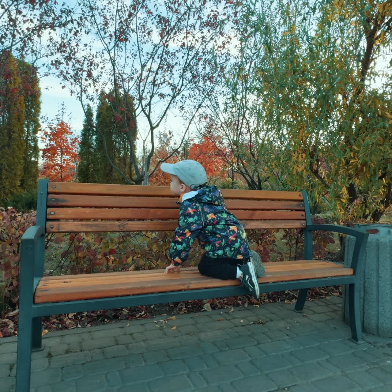 Lovely toddler runs by the park in autumn. Kid sits down on the bench and his grandpa gives him a magazine
