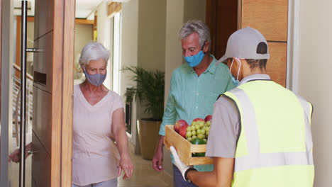 delivery man delivering groceries to senior caucasian couple wearing face masks at home