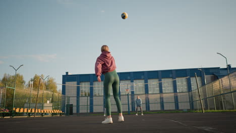 beginner volleyball player slamming ball incorrectly with two ladies watching from the distance, building visible in the background, showing playful and learning sports moment