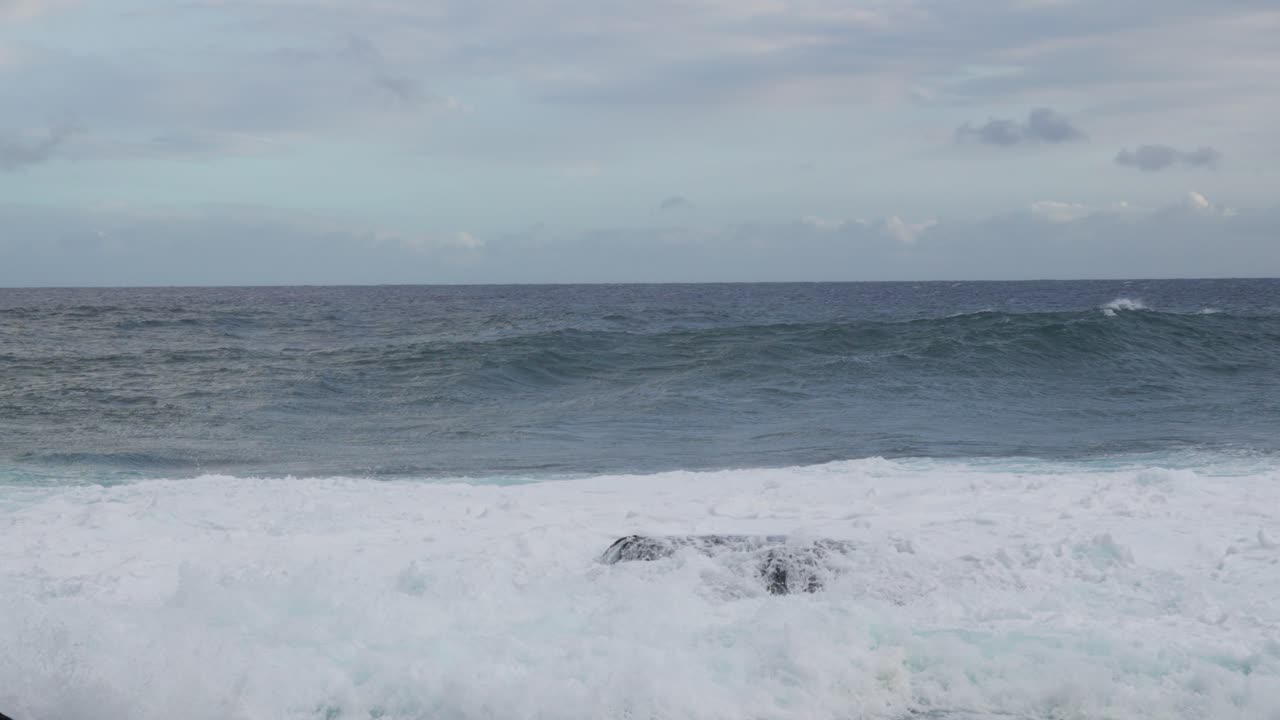 A rising set collides with a reef rock and erupts into white spray, churning foam across the frame while gray skies and restless swell convey raw, wind-driven energy