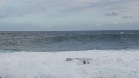 a rising set collides with a reef rock and erupts into white spray, churning foam across the frame while gray skies and restless swell convey raw, wind-driven energy